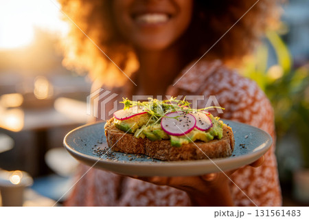 Close-up of a woman holding a sandwich with toasted bread sliced avocado green herbs and red radishes seasoned with pepper flakes. Close-up of a woman holding a sandwich with toasted bread sliced avocado green herbs and red radishes seasoned with pepper flakes. 131561483