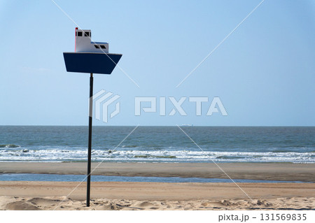 Wooden model of boat on pole on sandy beach with real big cargo ship in background, sunny day 131569835