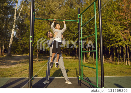 Young father helping a child boy to pull up himself on horizontal bar in summer park outdoors. 131569893