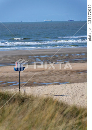 Wooden model of boat on pole on sandy beach with real big cargo ship in background, sunny day 131572039