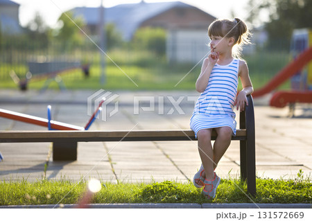 Cute young girl in short dress sitting alone outdoors on playground bench on sunny summer day. 131572669