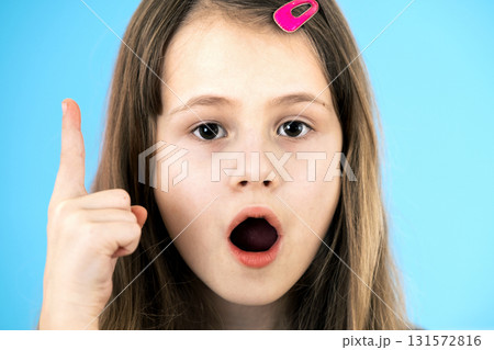Close up portrait of a child school girl holding up point finger in I have an idea gesture isolated on blue background. 131572816