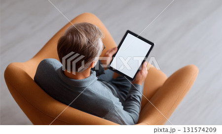 Calm man sitting in comfortable armchair at home, focused on reading digital tablet with blank screen. person enjoys leisure time relaxing with modern technology 131574209