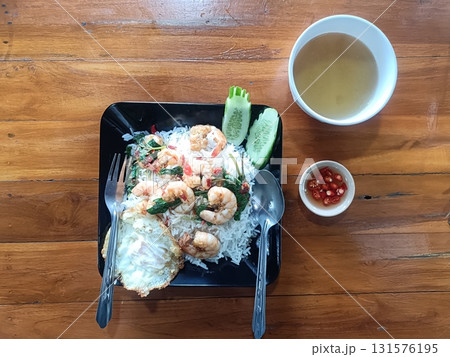 Stir fried shrimp with basil served over white rice with fried egg, cucumber slices, chili sauce and clear soup on wooden table, appetizing casual meal 131576195