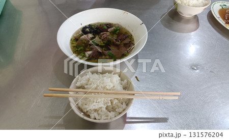 Bowl of clear beef soup with herbs and sliced meat served with steamed rice and chopsticks on stainless table, warm and savory meal Bowl of clear beef soup with herbs and sliced meat served with steamed rice and chopsticks on stainless table, warm and savory meal 131576204