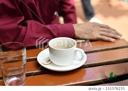 White coffee cup on saucer with used spoon on wooden table near elderly hand in red shirt conveying quiet morning contemplation 131576235