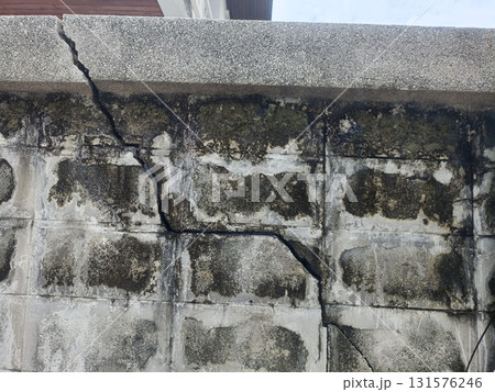 Cracked concrete wall with weathered stains and moss, vertical and diagonal fissure revealing structural damage and textured aged surface conveying decay and neglect Cracked concrete wall with weathered stains and moss, vertical and diagonal fissure revealing structural damage and textured aged surface conveying decay and neglect 131576246