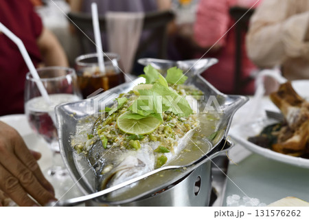 Steamed whole fish with lime, garlic and herb garnish served on metal platter in busy restaurant, fresh seafood and vibrant citrus aroma 131576262