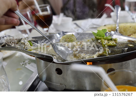 Steaming whole fish in savory broth on tabletop grill with spoon serving, herb garnish and communal dining atmosphere Steaming whole fish in savory broth on tabletop grill with spoon serving, herb garnish and communal dining atmosphere 131576267