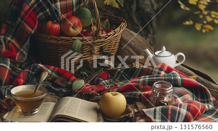 Autumn picnic setup with apples, tea, and book on plaid blanket, surrounded by basket of apples and teapot, creating cozy fall atmosphere 131576560
