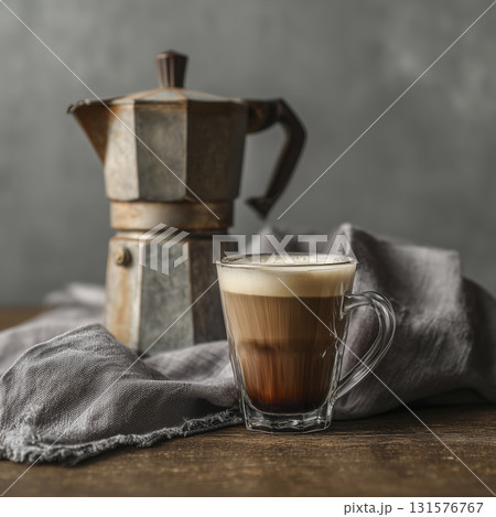 Glass cup of layered coffee with cream top and vintage moka pot background, placed rustic wooden surface with gray cloth, evokes cozy and warm Glass cup of layered coffee with cream top and vintage moka pot background, placed rustic wooden surface with gray cloth, evokes cozy and warm 131576767