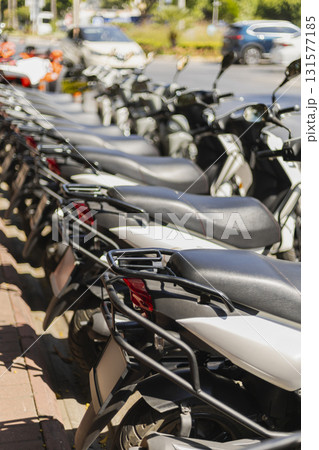 A row of parked scooters awaits riders along a busy city street. The sun shines brightly, highlighting the sleek design and vibrant colors of the vehicles 131577185
