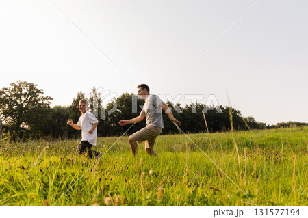 A father and son share a delightful moment in nature, running freely through a lush green field under a clear sky, surrounded by the beauty of trees and sunlight A father and son share a delightful moment in nature, running freely through a lush green field under a clear sky, surrounded by the beauty of trees and sunlight 131577194
