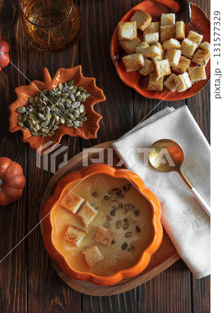 Creamy pumpkin soup with croutons and seeds, served in a pumpkin-shaped bowl on dark wooden table. Top view Creamy pumpkin soup with croutons and seeds, served in a pumpkin-shaped bowl on dark wooden table. Top view 131577329