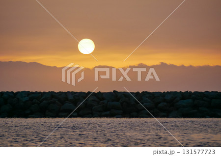 A serene sunset over a calm sea, with a large sun setting behind cloudy mountains. 131577723