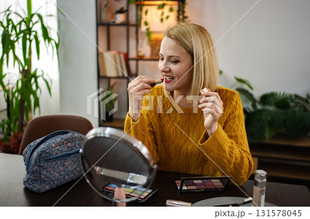Young woman applying makeup at a cozy table in a bright indoor space full of plants and personal touches while smiling happily Young woman applying makeup at a cozy table in a bright indoor space full of plants and personal touches while smiling happily 131578045