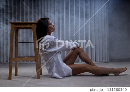 A beautiful woman sits gracefully on a soft floor wearing a white men's shirt. She leans against a wooden stool surrounded by gentle lighting and a serene atmosphere creating an artistic vibe. A beautiful woman sits gracefully on a soft floor wearing a white men's shirt. She leans against a wooden stool surrounded by gentle lighting and a serene atmosphere creating an artistic vibe. 131580540