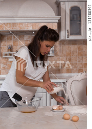 pregnant woman is whisking eggs with concentration. Flour dusts the countertop showing her involvement in cooking. The atmosphere feels inviting and nurturing. pregnant woman is whisking eggs with concentration. Flour dusts the countertop showing her involvement in cooking. The atmosphere feels inviting and nurturing. 131580562