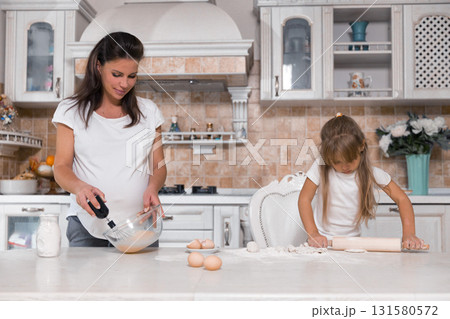 pregnant woman mixes batter in a bowl while her daughter rolls dough with a rolling pin. They are enjoying quality time together in their bright kitchen on a cheerful afternoon. pregnant woman mixes batter in a bowl while her daughter rolls dough with a rolling pin. They are enjoying quality time together in their bright kitchen on a cheerful afternoon. 131580572