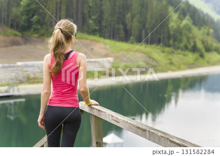 Back view of a young sportive woman in fitness clothes standing outdoors near big lake in mountains enjoying nature view. Back view of a young sportive woman in fitness clothes standing outdoors near big lake in mountains enjoying nature view. 131582284