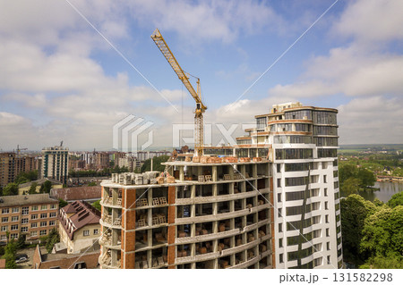 Apartment or office tall building under construction. Brick walls, glass windows, scaffolding and concrete support pillars. Tower crane on bright blue sky copy space background. Apartment or office tall building under construction. Brick walls, glass windows, scaffolding and concrete support pillars. Tower crane on bright blue sky copy space background. 131582298