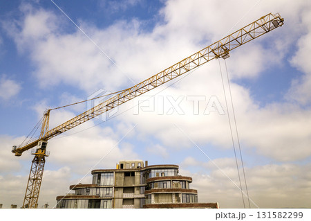 Apartment or office tall building under construction. Brick walls, glass windows, scaffolding and concrete support pillars. Tower crane on bright blue sky copy space background. Apartment or office tall building under construction. Brick walls, glass windows, scaffolding and concrete support pillars. Tower crane on bright blue sky copy space background. 131582299
