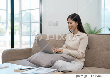 Young Asian woman happily working on a laptop while sitting on a couch at home, enjoying a productive day. 131586022