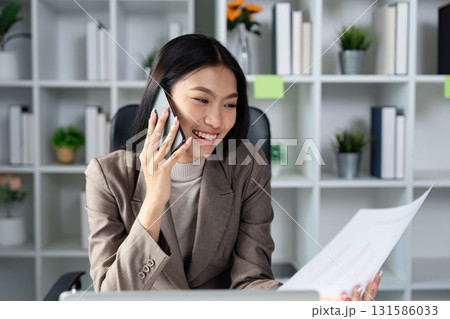 Smiling businesswoman on a phone call while reviewing financial documents in an office Smiling businesswoman on a phone call while reviewing financial documents in an office 131586033