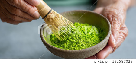 Elderly hand holding bamboo whisk mixing vibrant green matcha powder in rustic ceramic bowl, symbolizing longevity, tradition, wellness, and mindful tea preparation in serene setting Elderly hand holding bamboo whisk mixing vibrant green matcha powder in rustic ceramic bowl, symbolizing longevity, tradition, wellness, and mindful tea preparation in serene setting 131587025