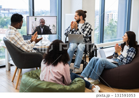 Diverse group of colleagues including man in wheelchair and a woman engaging in virtual meeting with African male speaker on screen, demonstrating teamwork, inclusivity, online collaboration. Diverse group of colleagues including man in wheelchair and a woman engaging in virtual meeting with African male speaker on screen, demonstrating teamwork, inclusivity, online collaboration. 131591053
