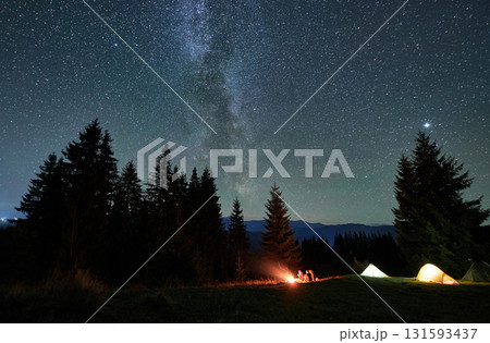Night camping in mountains. Amidst forested mountain landscape, two tents glow warmly beside campfire under brilliant, star-filled sky. Silhouettes of tourists resting at campsite under Milky Way. 131593437