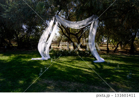 Outdoor wedding ceremony with a white fabric arch among trees and on grass Outdoor wedding ceremony with a white fabric arch among trees and on grass 131601411