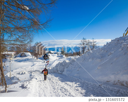 スキー板を担いでペンション街の雪道を歩くスキーヤー (新潟県、斑尾高原) 131605572