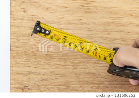 Hand holding tape measure yellow tape metal hook wood board measurement tool DIY construction carpentry precision closeup view shows accurate scale for project planning and careful craftsmanship 131606252