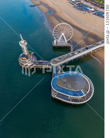 Aerial view of ocean pier with Ferris wheel, glass roofed building, and spiral tower set against busy beach and deep blue water, highlighting vibrant coastal recreation. 131606780