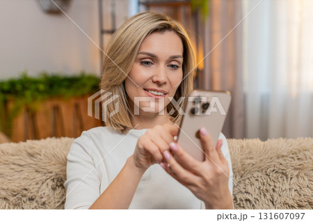 Adult woman thoughtfully writing text message on smartphone at home, smiling while thinking quietly Adult woman thoughtfully writing text message on smartphone at home, smiling while thinking quietly 131607097