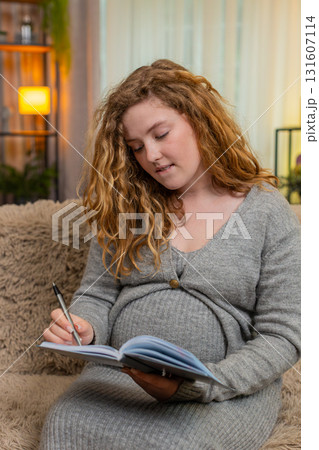 Pregnant young woman writing in diary to do list at home, thoughtful and focused, future planning 131607114