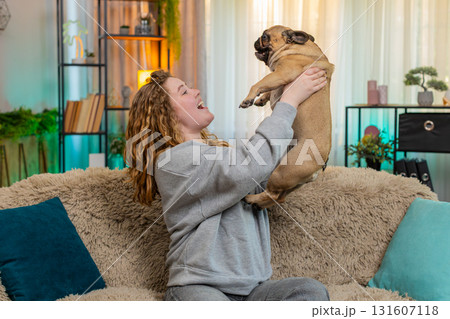 Young woman lifts pug dog with joy on sofa, celebrating success and sharing happy cheerful emotion 131607118