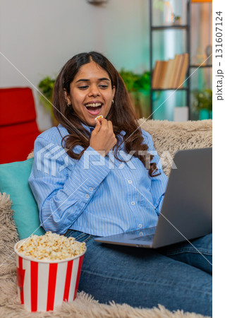 Indian woman watching movie with popcorn smiling at laptop screen on home sofa during cozy moment 131607124