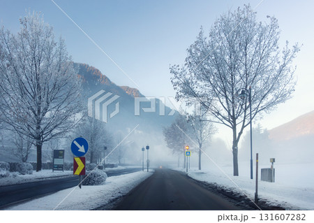 Scenic quiet winter road frosty valley covered mist fog and snow. Bare trees and distant mountains glow softly under the pale morning light. Alpine mountain village winter morning sunrise Scenic quiet winter road frosty valley covered mist fog and snow. Bare trees and distant mountains glow softly under the pale morning light. Alpine mountain village winter morning sunrise 131607282