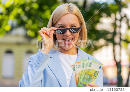 Happy rich mature business woman putting on thug life sunglasses showing euro money on city street 131607284