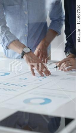Business people are examining financial data, pointing at charts and graphs spread out on a table during a collaborative analysis session in a modern office, close up vertical view Business people are examining financial data, pointing at charts and graphs spread out on a table during a collaborative analysis session in a modern office, close up vertical view 131607516