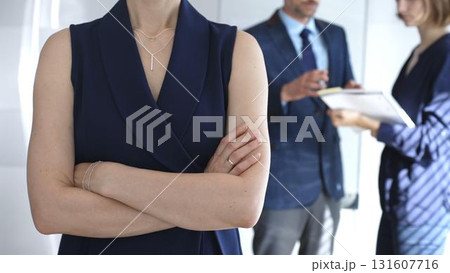 Professional businesswoman stands confidently with arms crossed in sharp focus as colleagues interact behind her during an office meeting, symbolizing leadership and corporate success Professional businesswoman stands confidently with arms crossed in sharp focus as colleagues interact behind her during an office meeting, symbolizing leadership and corporate success 131607716