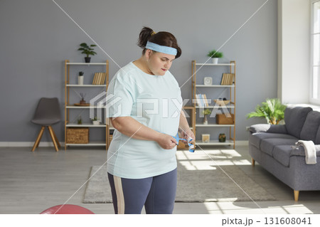 Fat woman wearing sportswear measure waist with measuring tape standing at home. Fat woman wearing sportswear measure waist with measuring tape standing at home. 131608041