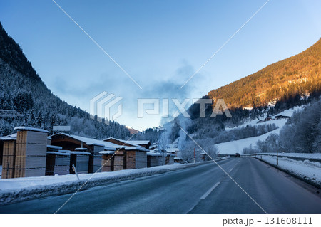 Scenic quiet winter road winds through a mountain valley beside a lumber yard. Smoke rises from village houses as golden sunlight warms forested slopes above the frozen landscape. Scenic quiet winter road winds through a mountain valley beside a lumber yard. Smoke rises from village houses as golden sunlight warms forested slopes above the frozen landscape. 131608111