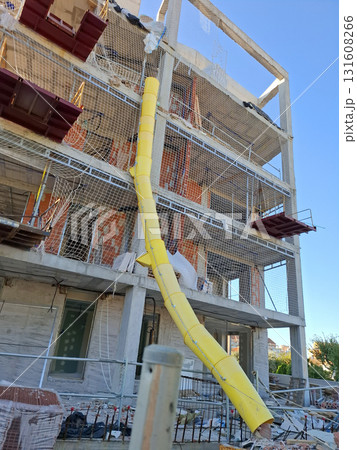 Workers are constructing a multi-story building with bright yellow debris netting and a chute attached to the roof for safely disposing of materials. Workers are constructing a multi-story building with bright yellow debris netting and a chute attached to the roof for safely disposing of materials. 131608266