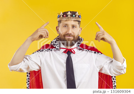 Portrait of young confident man in suit with crown looking at camera on yellow background. 131609304