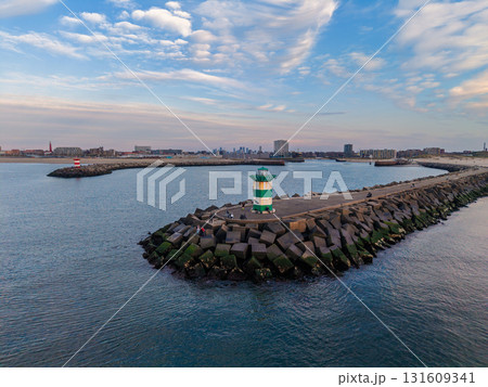 Aerial view of coastal breakwater with green striped lighthouse, angular concrete blocks, and distant cityscape blending maritime infrastructure with urban backdrop under scenic skies. Aerial view of coastal breakwater with green striped lighthouse, angular concrete blocks, and distant cityscape blending maritime infrastructure with urban backdrop under scenic skies. 131609341