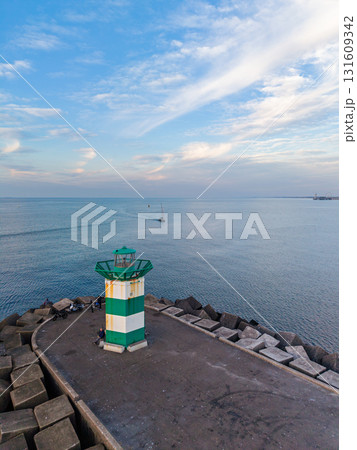 Aerial view of concrete pier with green-striped lighthouse and wave breakers, sailboat in distance capturing serene maritime atmosphere under soft, streaked clouds. Aerial view of concrete pier with green-striped lighthouse and wave breakers, sailboat in distance capturing serene maritime atmosphere under soft, streaked clouds. 131609342