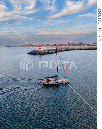 Aerial view of sailboat approaching curved breakwater with red striped lighthouse, calm sea, and urban skyline capturing serene maritime navigation and coastal city backdrop. 131609344